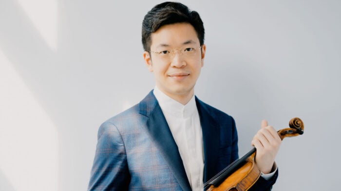 Paul Huang stands against a sunlit wall, wearing a blue sportcoat, holding a violin