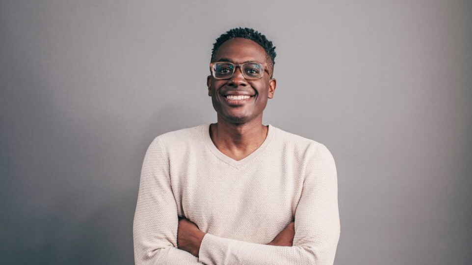 Headshot of composer Shawn E. Okpebholo, smiling with arms crossed in front of a grey background