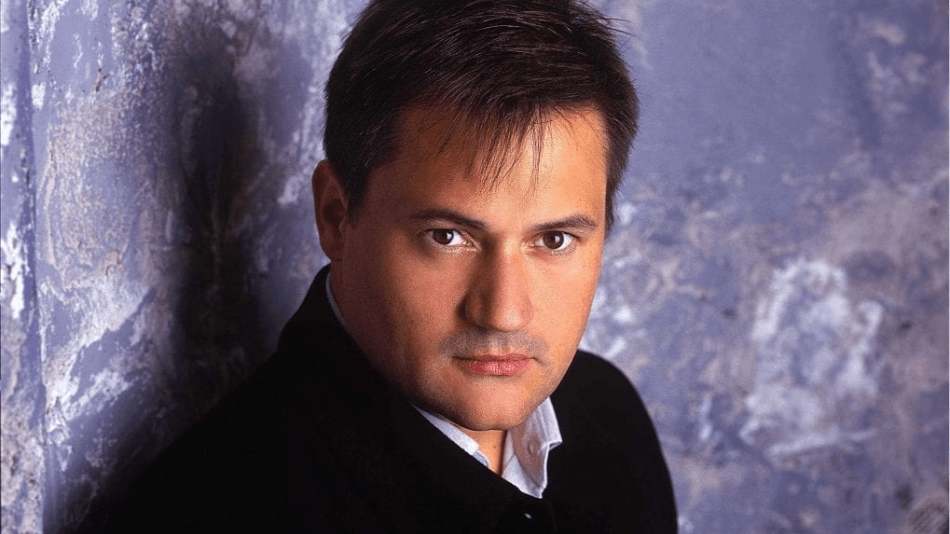Portrait of Paul Groves looking over his right shoulder directly into camera; dark jacket, white shirt, lavender colored background.