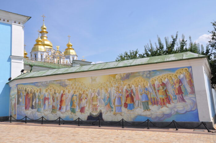 A fresco corwned and haloed archangels and angels standing on a cloud. A Baroque-Byzantine church with gold domes is in the background