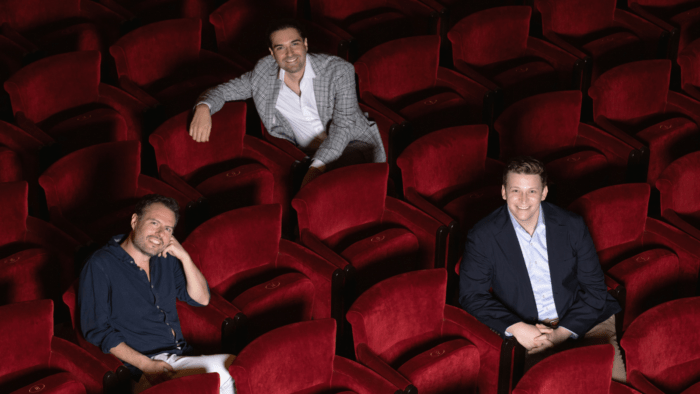 Raffaele Pe, Carlo Vistoli and Aryeh Nussbaum Cohen seated on the red upholstered seats of the auditorium of Teatro dell'Opera di Roma; smiling in business casual attire.