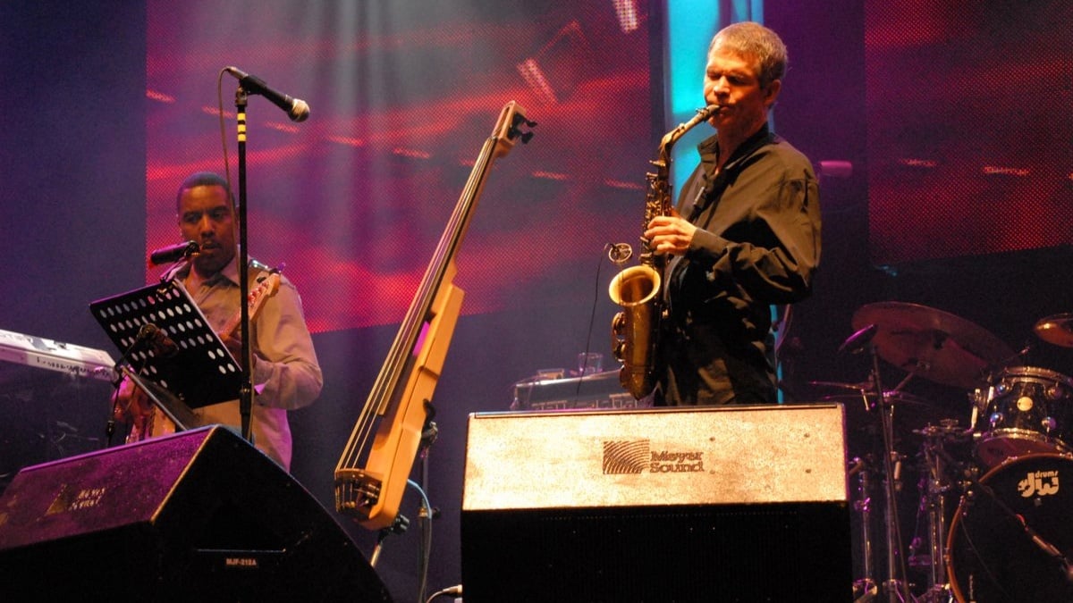 David Sanborn is absorbed as he plays a saxophone onstage; a bandmate holding a bass guitar looks on