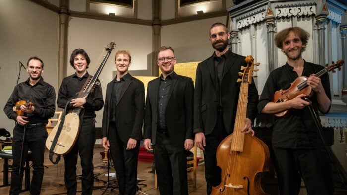 five male musicians, dressed in black suite with black shirts, holding a variety of early music stringed instruments .