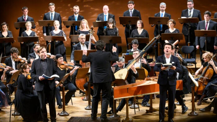Two male singers front an early music ensemble and a row of women singers and a row of male singers. A conductor faces the ensemble.