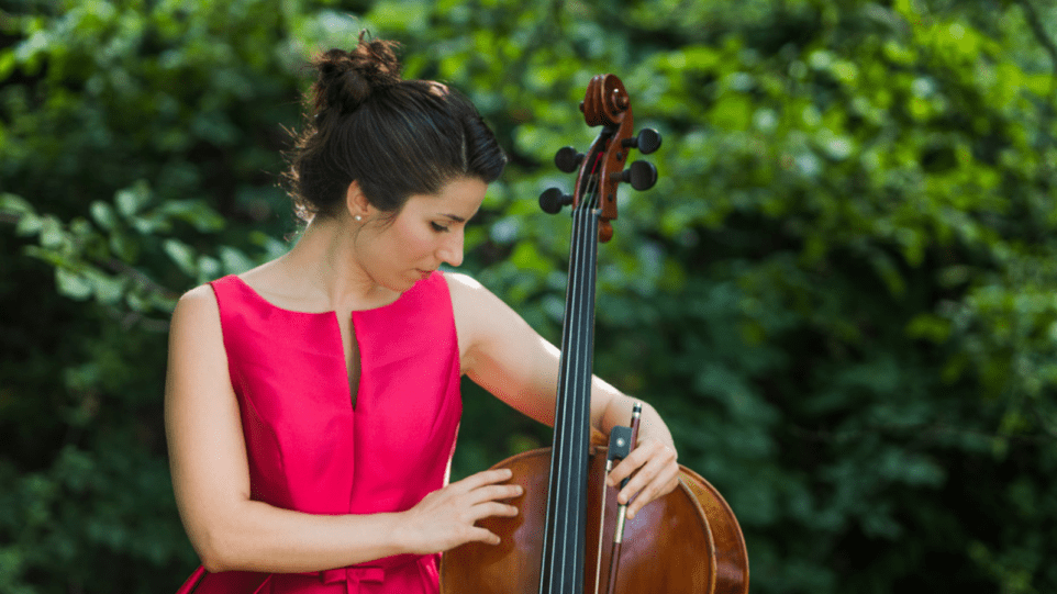 Composer and cellist Andrea Casarrubios poses with her cello by her side. She is looking down on the instrument with a soft expression and wearing a pink dress.