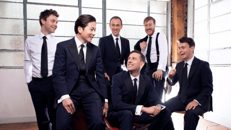 Portrait of the six gentlemen of the King's Singers in a casual pose in dark suits with white shirts, two with their jackets removed, in a room filled with natural light.