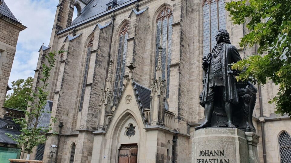 A statue of composer Johann Sebastian Bach, dressed in ealry 18th century German attire in front of a large Romanesque/Gothis church.