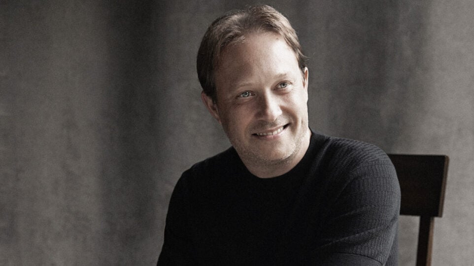 Ludovic Morlot, seated, wears a dark knit short sleeve shirt and grins against a studio backdrop