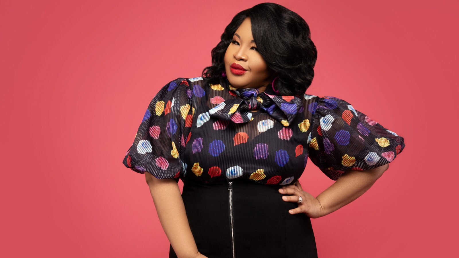 in a multicolored polka dot blouse and a black skirt, Karen Slack poses against a pink background