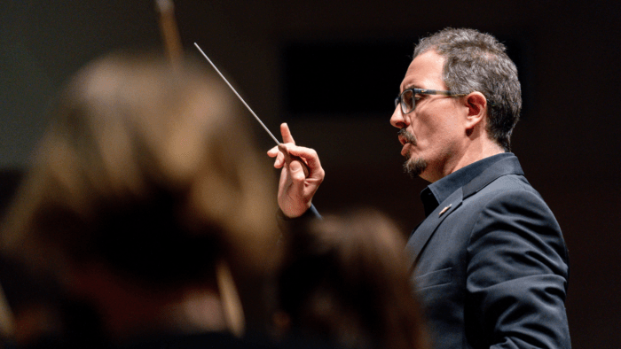 Profile of Emanuele Andrizzi conducting, baton in right hand gesturing; musician in soft focus in foreground.