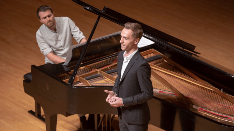 Bariotne Dominik Belavy standing in the crook of Steinway grand piano, wearing a dark suit, light shirt, no tie; pianist Daniel Gerzenberg at the keyboard, in shirt sleeves, rolled to elbow.