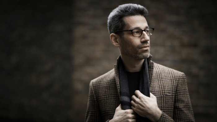 A photo of Jonathan Biss, a white male in his 40s, looking off to the right of the camera. He is set against a dark greyish-brown background, wearing a tweed coat, gray scarf, and black glasses.