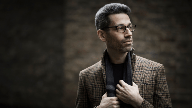 A photo of Jonathan Biss, a white male in his 40s, looking off to the right of the camera. He is set against a dark greyish-brown background, wearing a tweed coat, gray scarf, and black glasses.