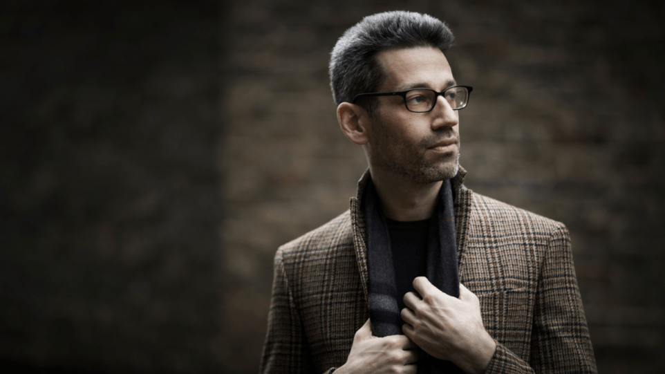 A photo of Jonathan Biss, a white male in his 40s, looking off to the right of the camera. He is set against a dark greyish-brown background, wearing a tweed coat, gray scarf, and black glasses.