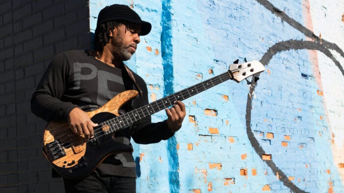 Victor Wooten plays bass guitar against a painted blue brick wall