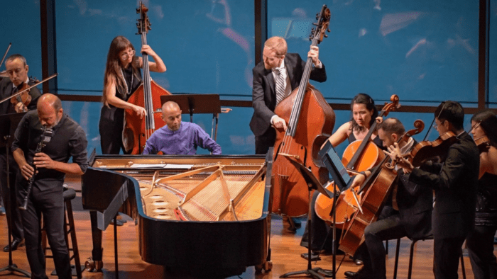 A group of orchestral musicians playing chamber music, dressed in black. In the center is a soloist in a purple shirt playing at the piano, surrounded by the ensemble.