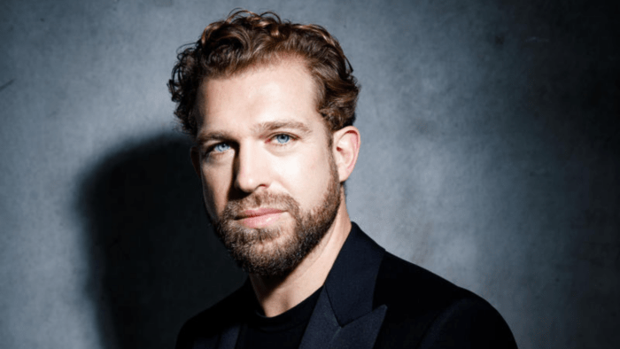 Portrait of Julian Prégardien in a dark suit, dark shirt, grey background, strong contrast created by lighting.