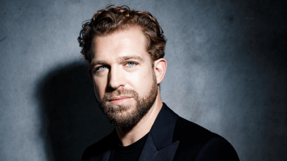 Portrait of Julian Prégardien in a dark suit, dark shirt, grey background, strong contrast created by lighting.