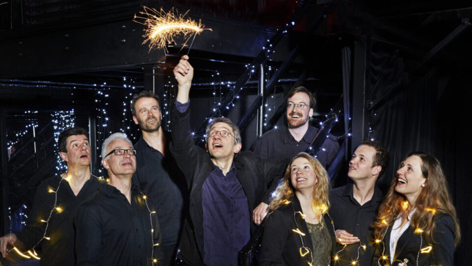 8 happy people adorned with lights on strings, on with a sparkler above his head