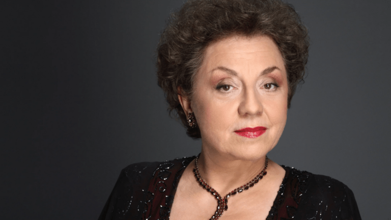 Portrait of Ewa Podleś in a beaded black dress and a dark beaded necklace facing the forward, dark background.