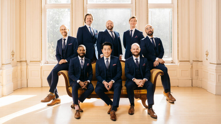 Eight men in suits sit on sofa in front of bright window