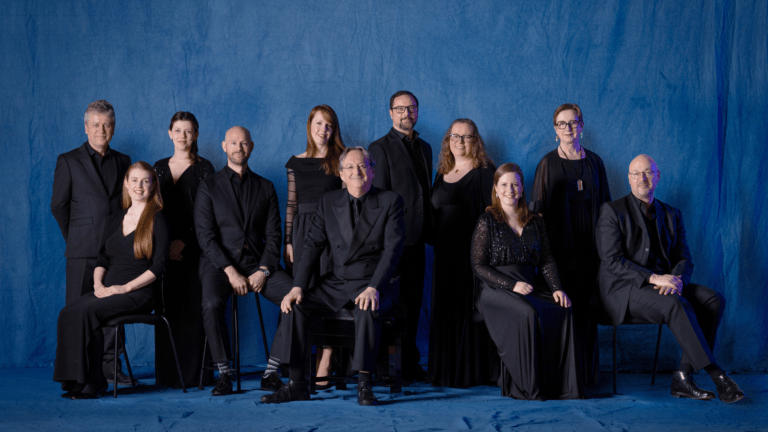 10 singers of the Tallis Scholars and conductor Peter Phillips, all in concert black, some seated, some standing, blue backdrop.