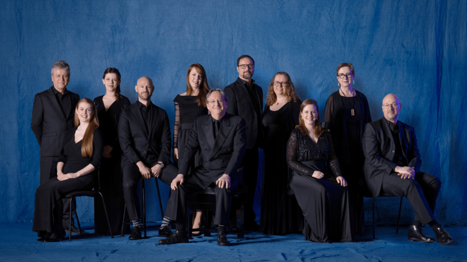 10 singers of the Tallis Scholars and conductor Peter Phillips, all in concert black, some seated, some standing, blue backdrop.