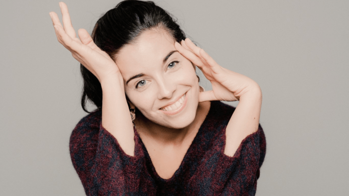 Whimsical portrait of Jodie Devos in brocade V-neck blouse, dark hair in a ponytail, head tilted towards the right, fingers of left hand touching her temple and jaw, palm of right hand touching her foreheard.