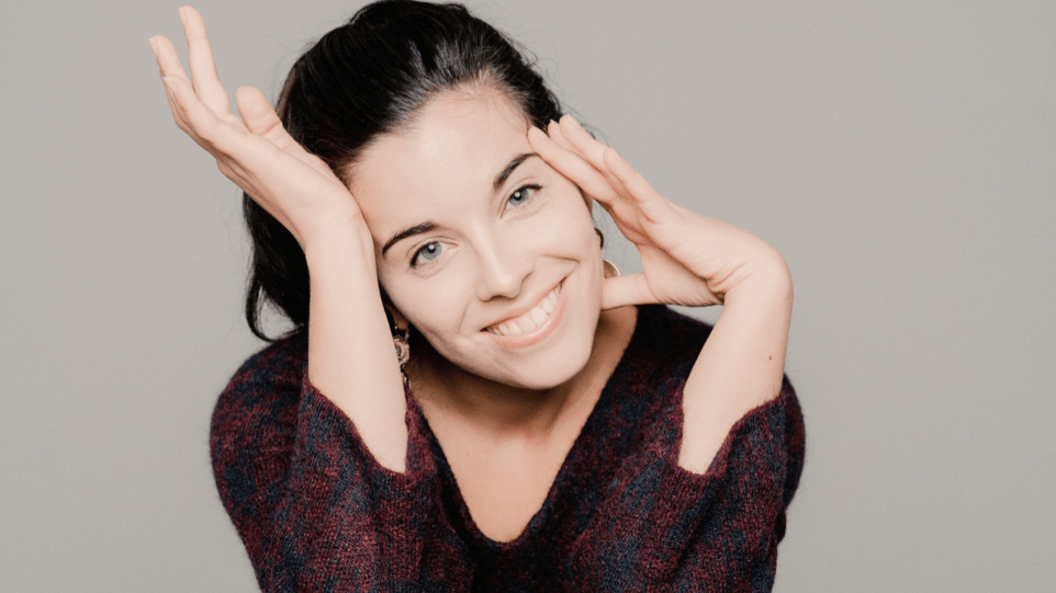 Whimsical portrait of Jodie Devos in brocade V-neck blouse, dark hair in a ponytail, head tilted towards the right, fingers of left hand touching her temple and jaw, palm of right hand touching her foreheard.