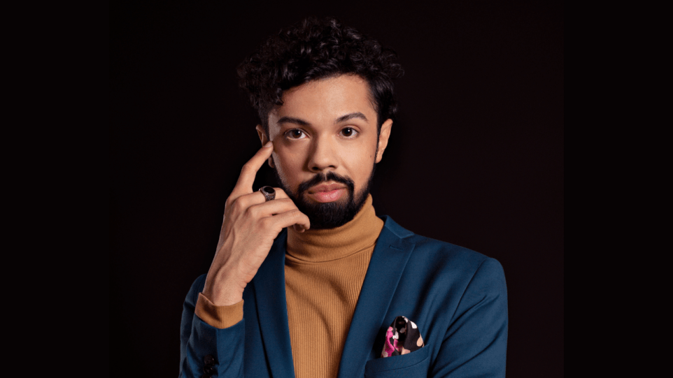 Bruno de Sá in a blue suit with a caramel colored turtleneck that very closely matches his skin tone; colorful print pocket square; pointing at his right eye ; black background