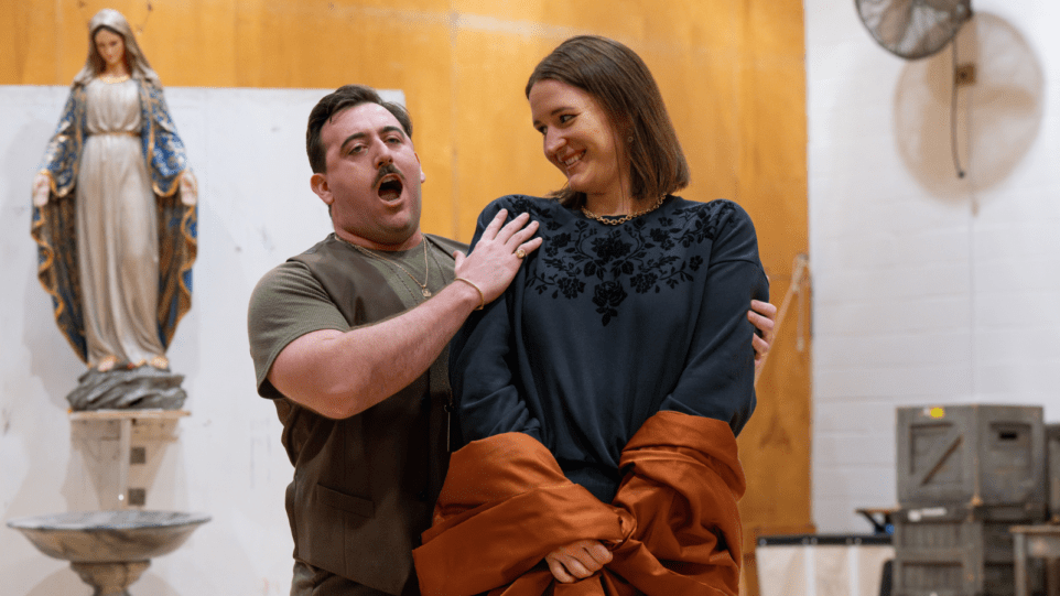 Freddie de Tommaso and Lise Davidsen in rehearsal for 'Tosca" at the Metropolitan Opera; wearing street clothes; De Tommaso singing with his right hand on Davidsen's right shoulder, his left hand reaching around her left arm; Davidsen smiling; statue of the Madonna behind them