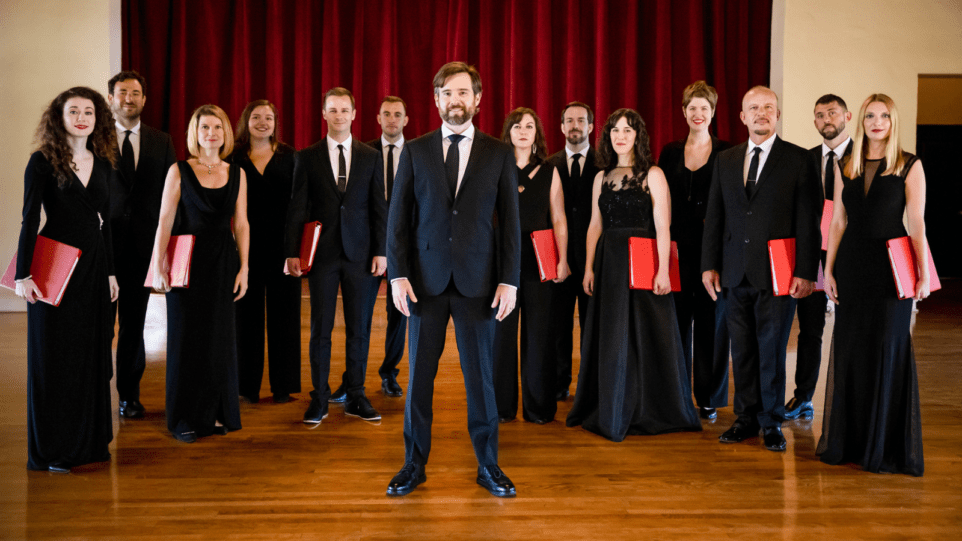 Group portrait of seven women and men of Seraphic Fire in concert black attire holding red choir folders; conductor Patrick Durpre Quigley in navy blue suit centered in image; hardwood floor, red curtain in background.