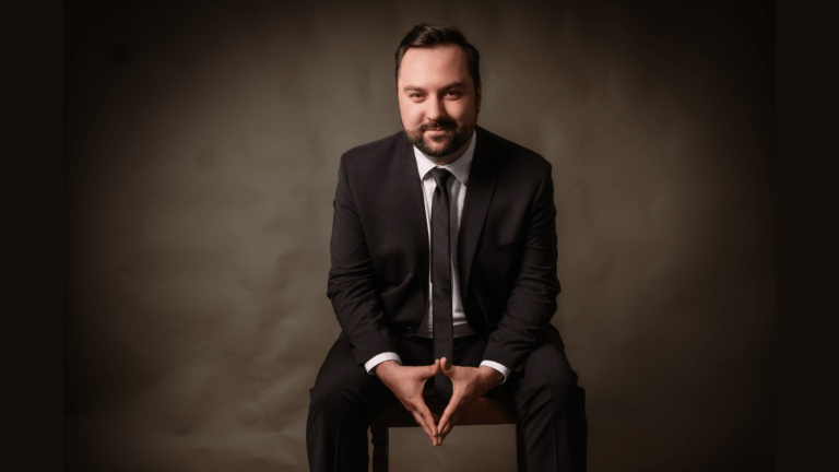 Professional studio portrait of Edward Vogel in dark suit, white shirt, dark tie, seated in front of a brown backdrop; elbows resting on knees, fingertips and thumbs of each hand touching.