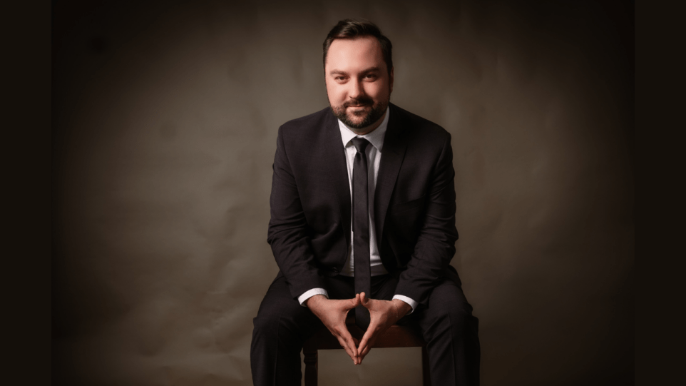 Professional studio portrait of Edward Vogel in dark suit, white shirt, dark tie, seated in front of a brown backdrop; elbows resting on knees, fingertips and thumbs of each hand touching.