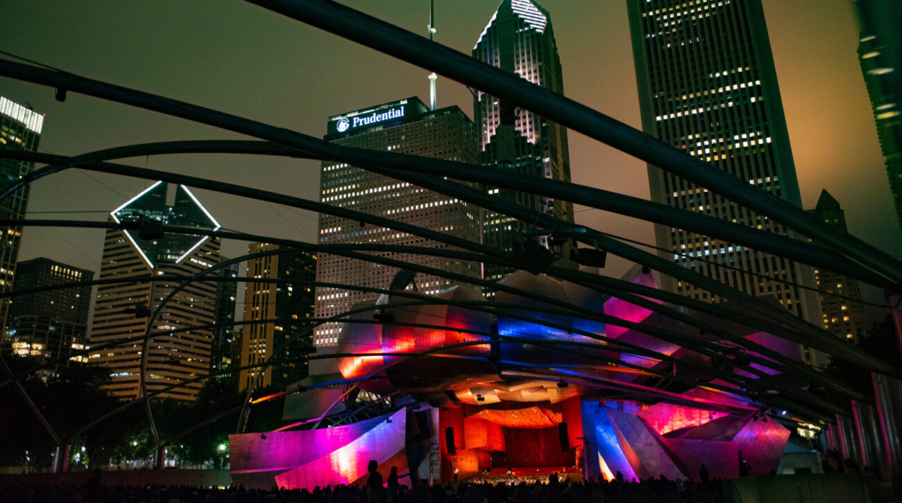 Nighttime at the Grant Park Music Festival as the public gazes upon the striking Pritzker Pavilion, lit in multicolor and with the Grant Park Orchestra onstage. The skyline in the background