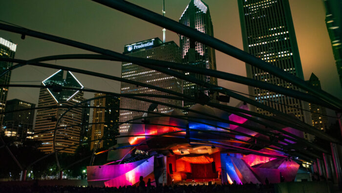 Nighttime at the Grant Park Music Festival as the public gazes upon the striking Pritzker Pavilion, lit in multicolor and with the Grant Park Orchestra onstage. The skyline in the background
