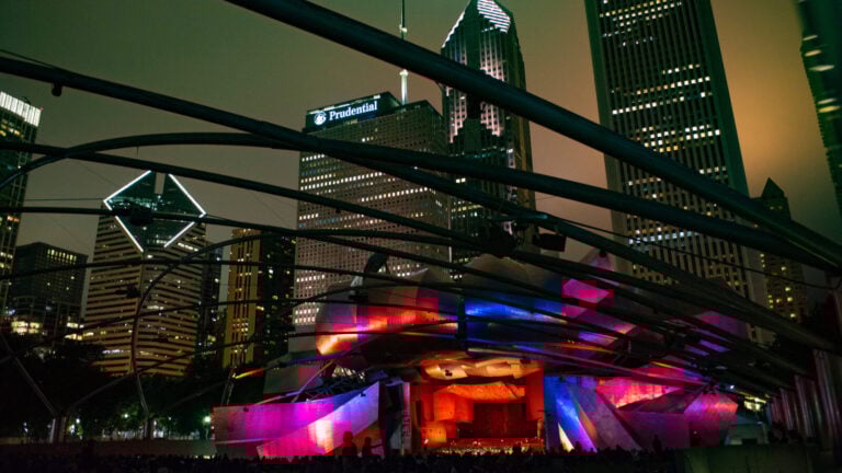 Nighttime at the Grant Park Music Festival as the public gazes upon the striking Pritzker Pavilion, lit in multicolor and with the Grant Park Orchestra onstage. The skyline in the background