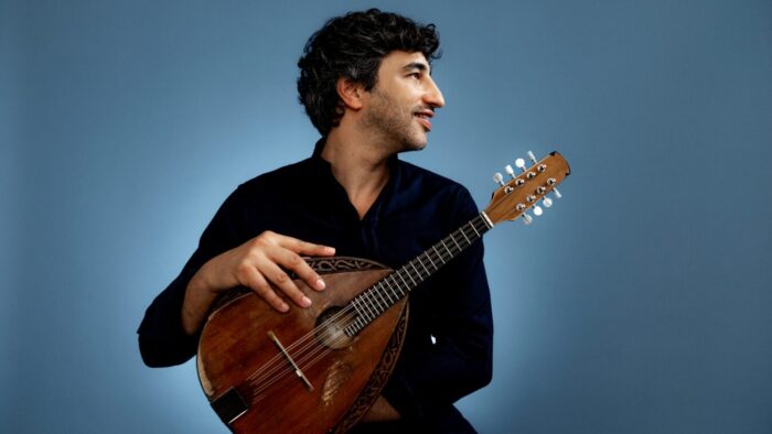 a young man looks over his let shoulder, his right arm holding a mandolin that rests on his knee