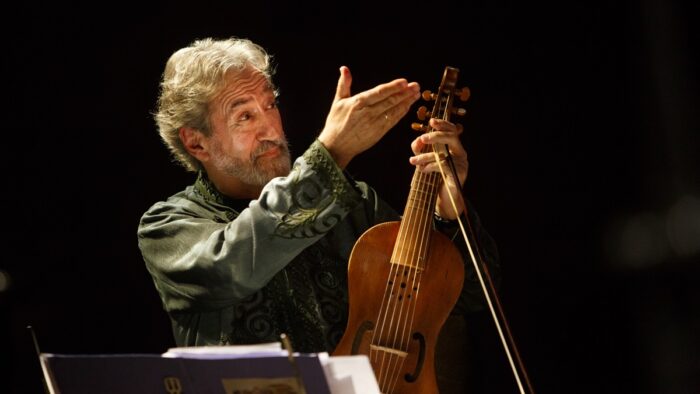 A man (Jordi Savall) holds a viol and bow in his left hand, his right hand outreachedm palm up