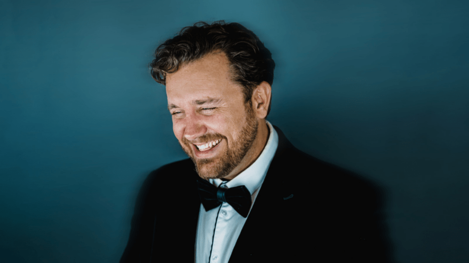 Portrait of Michael Spyres in a tuxedo, smiling so widely his eyes are shut, teal background.
