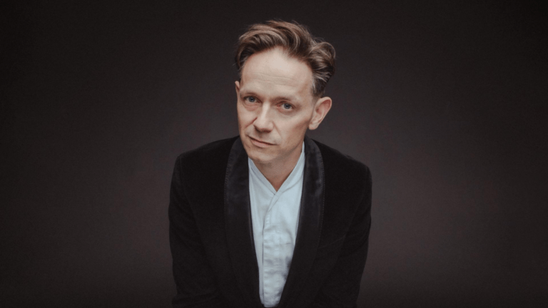 Portrait of Iestyn Davies in velvet tuxedo jacket, white shirt with band collar, no tie, leaning forward toward camera, dark background
