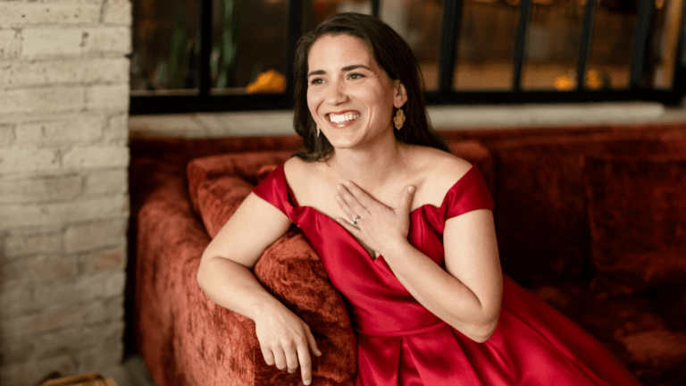 Joélle Harvey seated at the edge of a rust colored crushed velvet couch, in a red queen anne gown, her left hand touching her clavicle, smiling warmly.