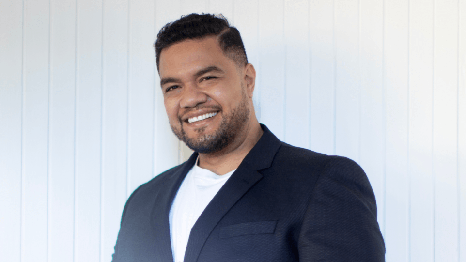 Portrait of Pene Pati, smiling, in dark suit over a white t-shirt; white background creating strong contrast with suit and skin tone.