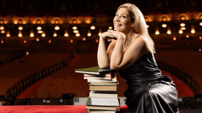 Sondra Radvanovsky in sleeveless black leather dress, elbows resting on a stack of opera scores, hands folded under chin, smiling, in a girlish pose, strawberry bonde hair, on stage of the Ardis Krainik at the Civic Opera House, empty auditorium out of focus in background.