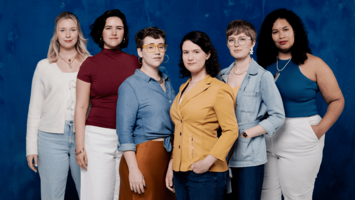 Symmetrical posed portrait of the six women of Lyyra in casual attire, lots of denim and white with some mustard, burgundy, and copper highlights in front of a ocean blue background.