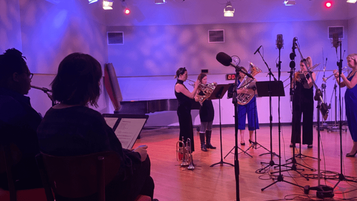 The all female brass quintet Seraph Brass recording a performance at WFMT's Levin Studio. The lights are mixed purple and blue. The hosts for Sounds Classical sit in two chairs to the left of the ensemble, observing them.