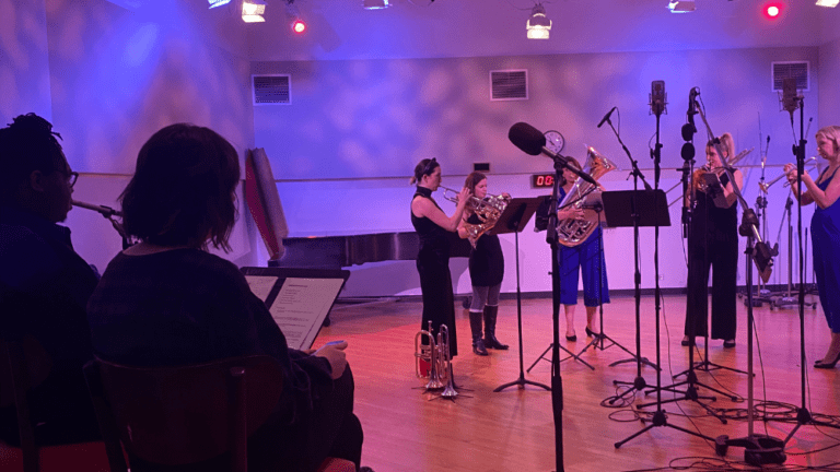 The all female brass quintet Seraph Brass recording a performance at WFMT's Levin Studio. The lights are mixed purple and blue. The hosts for Sounds Classical sit in two chairs to the left of the ensemble, observing them.