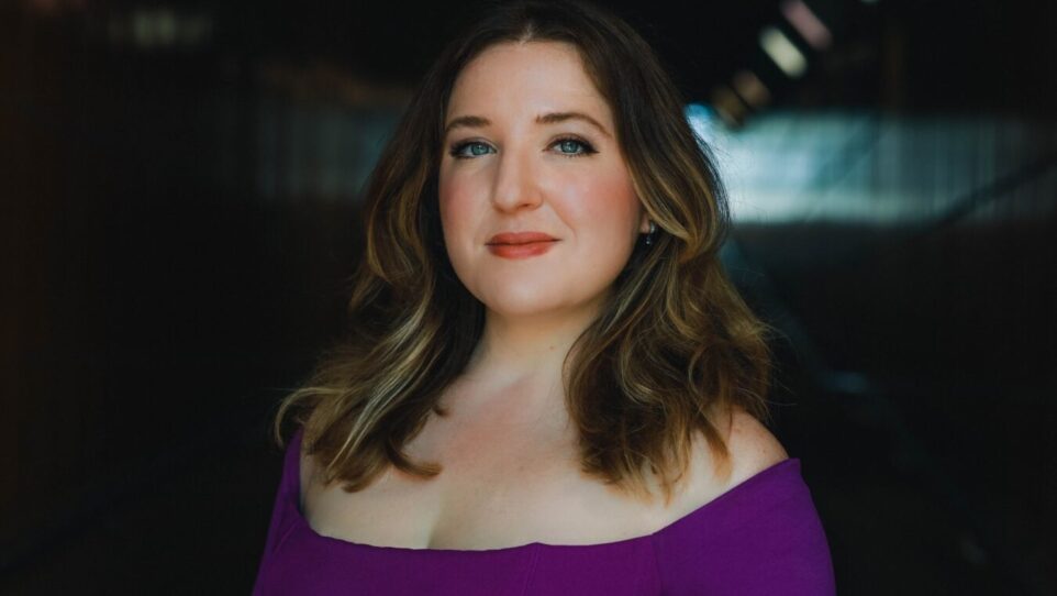 Portrait of Hannah De Priest in a purple, off-the-shoulder gown, wavy hair with blonde highlights; background is an obscured dark corridor.