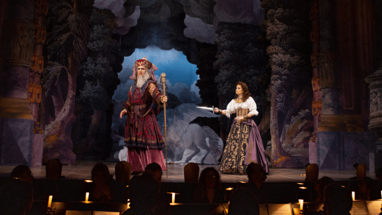 Jesse Blumberg and Emőke Baráth in full baroque costume in an opera performance, Baráth is pointing a sword at Blumberg who is turned a way from her. Orchestra visible in the bottom of the frame.