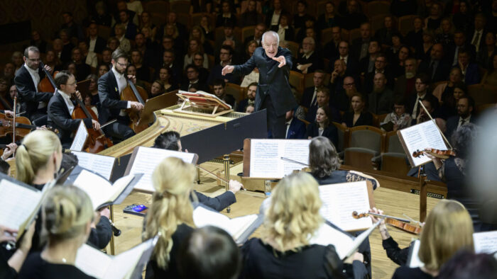 an older man conducts an orchestra with audience members behind him